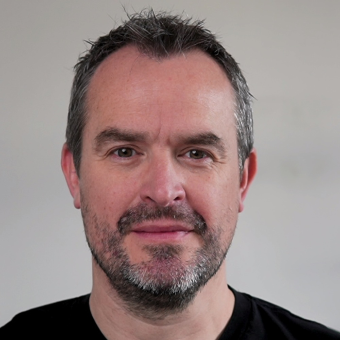 Photograph of Al, think Milk Tray Man, wearing a smile, a short beard and a black t-shirt, with blue eyes looking at the camera