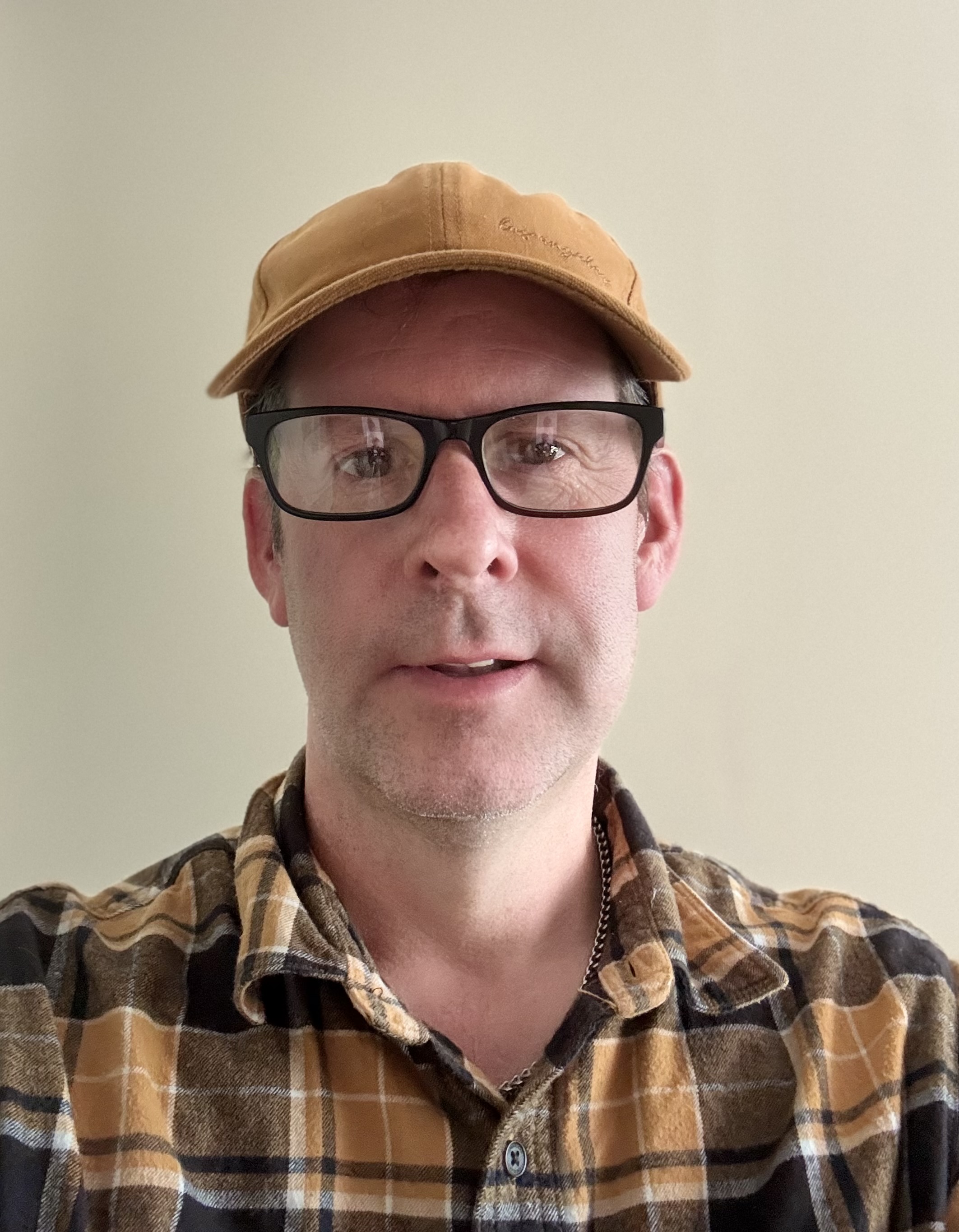 A photo of Lloydi facing the camera, wearing a tan baseball cap, dark-rimmed glasses, and a plaid shirt, looking thoughtful and ready for some in-depth accessibility conversations.