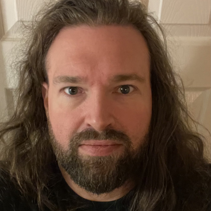 Photograph of Ian, looking ready to squash those accessibility bugs, with a beard and long wavy hair down to his shoulders, face on to the camera