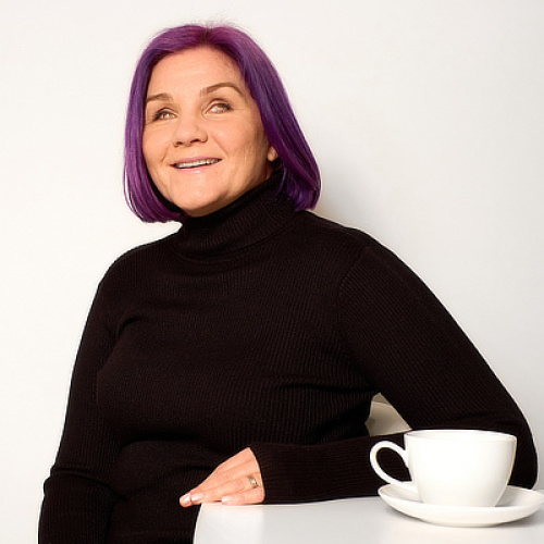 Photograph of Léonie, our purple-haired maverick, wearing a black polo neck, sitting at a table with a cup of tea, grinning at the camera