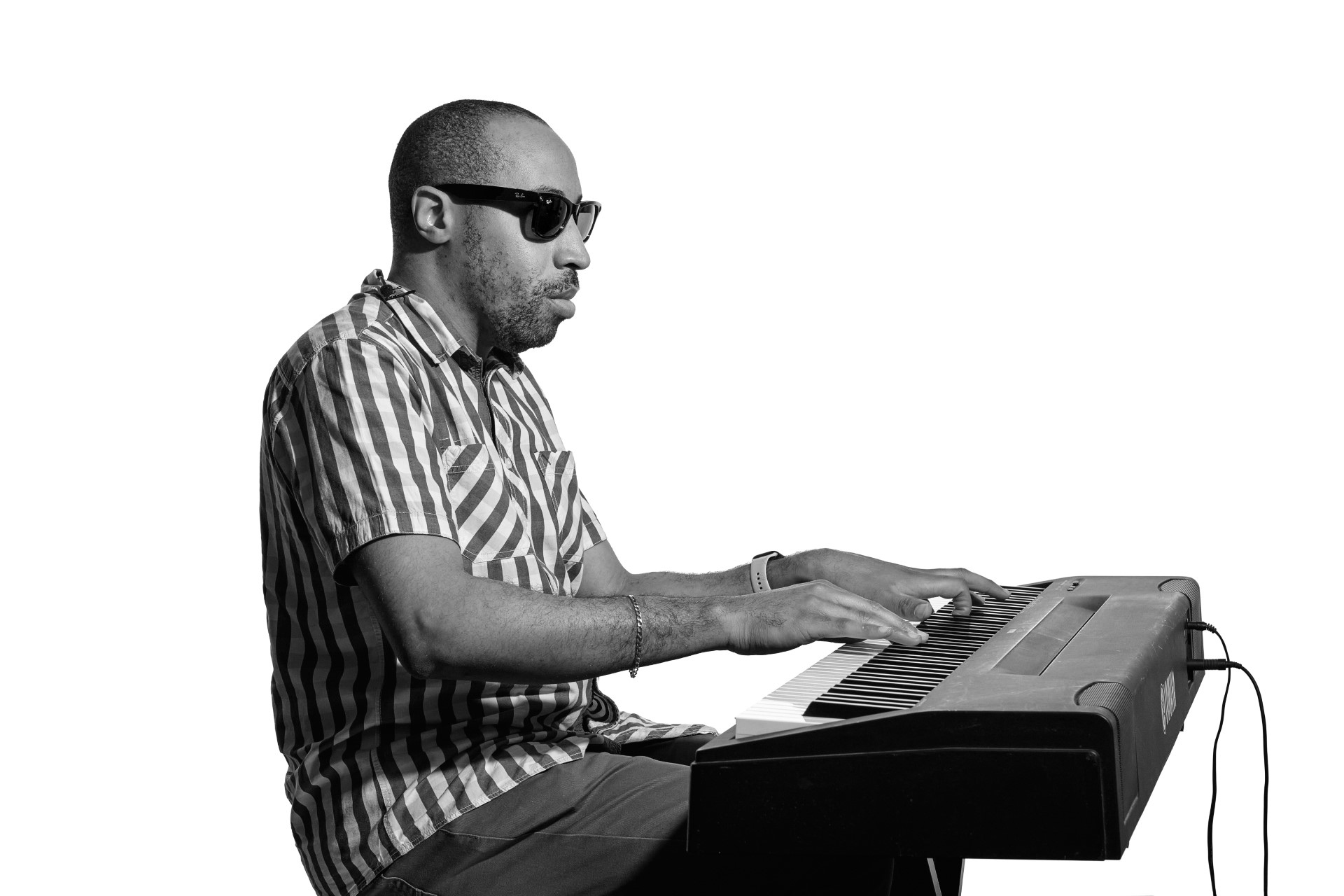 A black and white photo of Andre playing an electronic keyboard sitting side on. Andre is a younger black man and wears a striped shirt and Ray-Ban Meta Smart Glasses