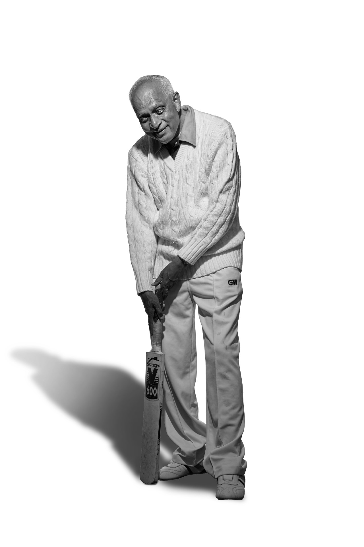 A black and white photo of Hasmukh, a man of South Asian heritage. He is wearing his cricket whites smiling as he stands ready to bat