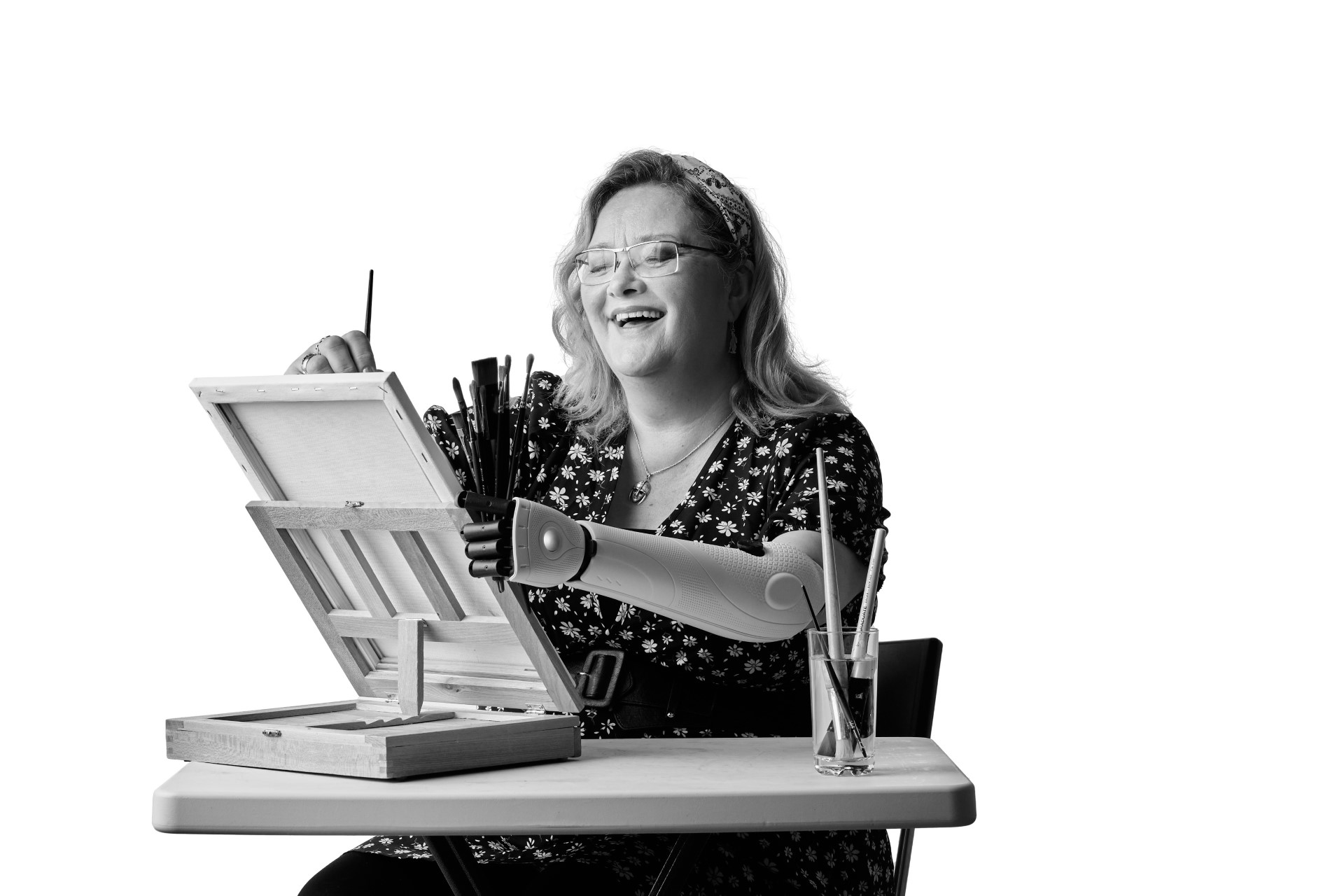 A black and white photo of Bev, a white woman in her 50s with blond hair scooped back in a hairband. She is sitting in front of a canvas laughing while she paints with her right hand and holds the canvas and some paint brushes in her prosthetic hand and arm