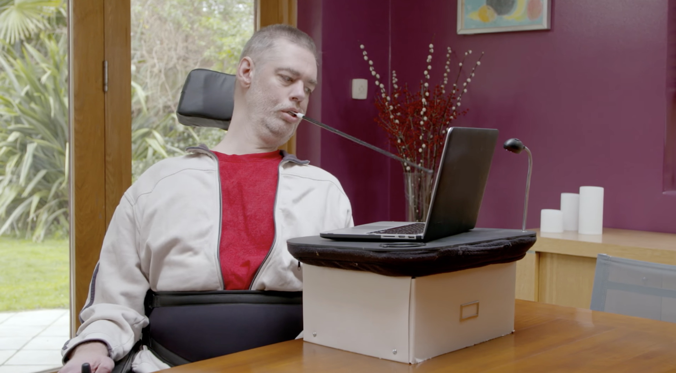 A man in a wheelchair at his desk, holding a pointing device with his mouth and using the keyboard on his laptop