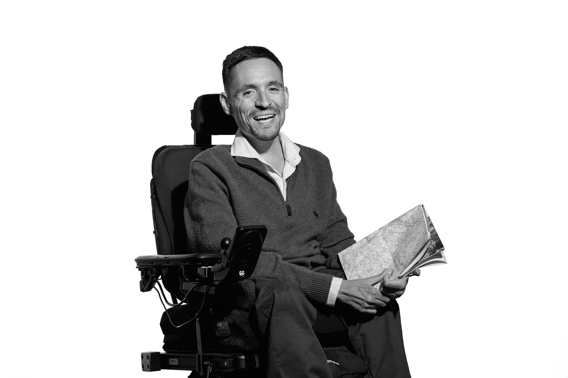 A black and white photo of Josh, a younger white man with short brown hair. He is sitting in his wheelchair as he laughs at the camera holding an ordnance survey map