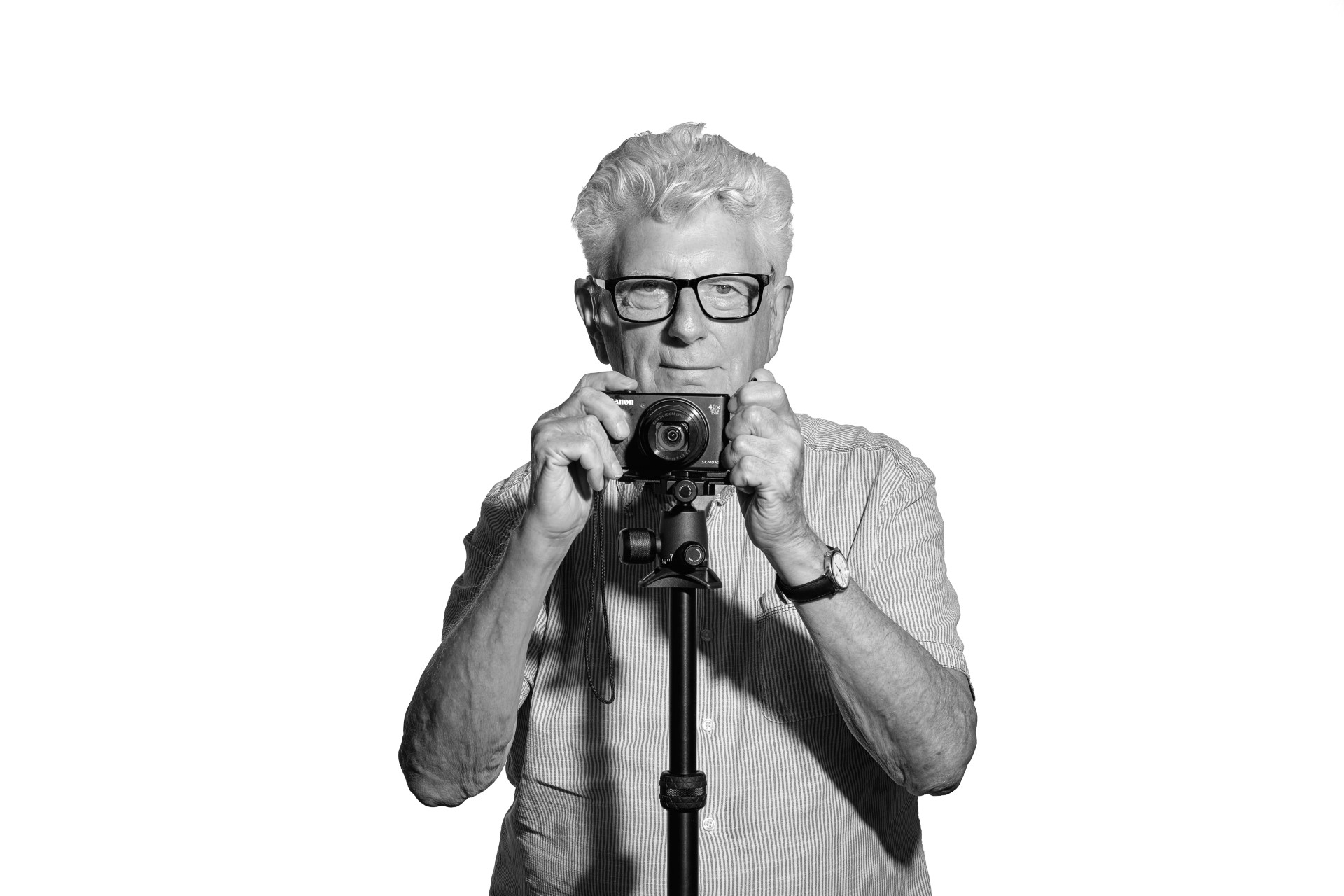 A black and white photo of Steve, an older white man with thick white hair. He is standing facing into the camera while he holds his book or poetry and photos open in front of him