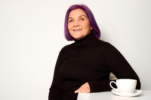 Photograph of Léonie, our purple-haired maverick, wearing a black polo neck, sitting at a table with a cup of tea, grinning at the camera