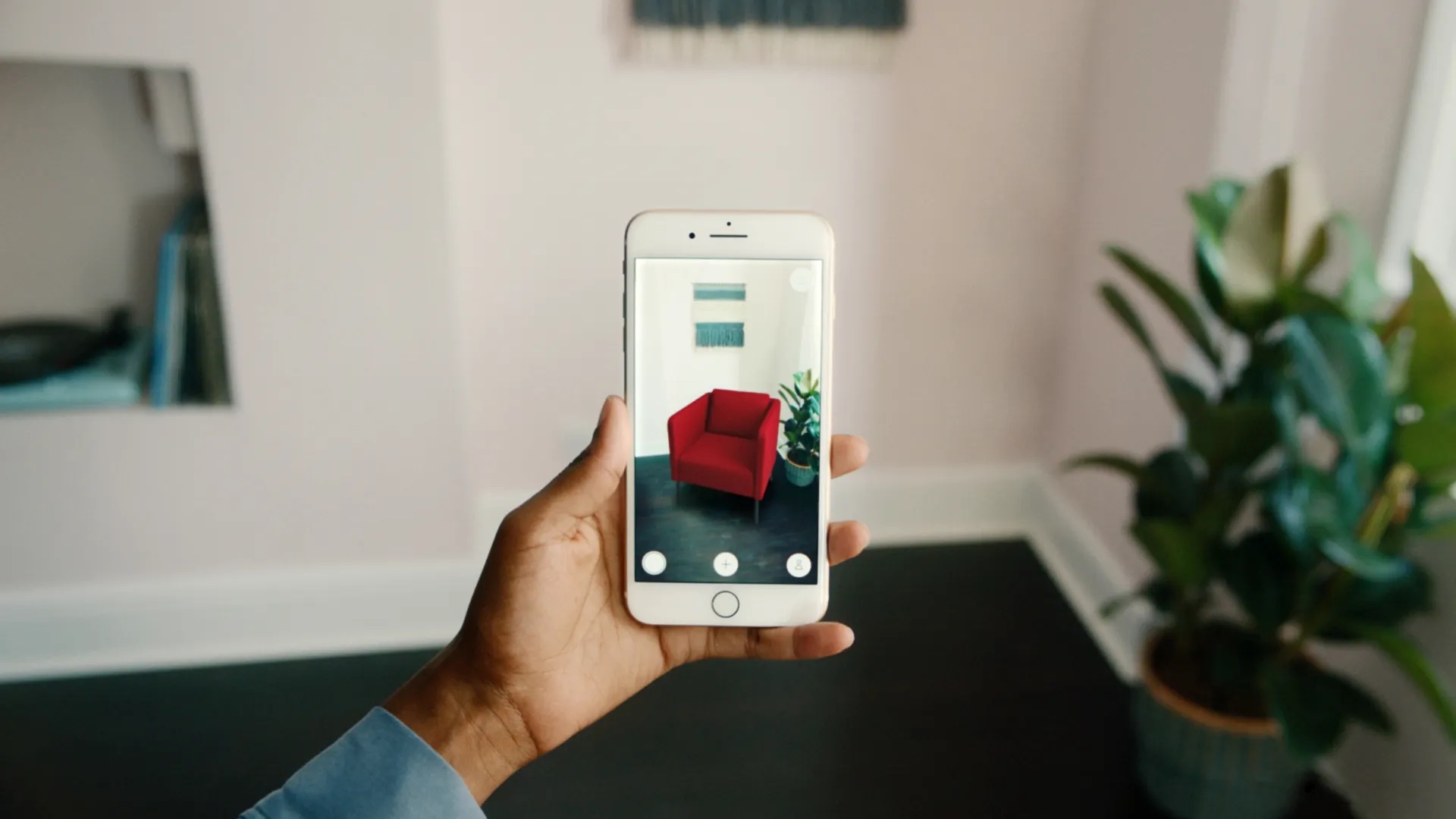 A person's arm holding out their mobile device, pointing at an empty corner of their room. On their device the camera feed is displaying the room, with a 3D model of an armchair visible in the empty space.