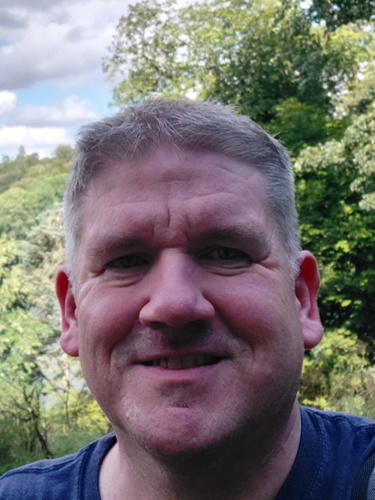 Dean, smiling in a close-up at the camera with a leafy green background behind him