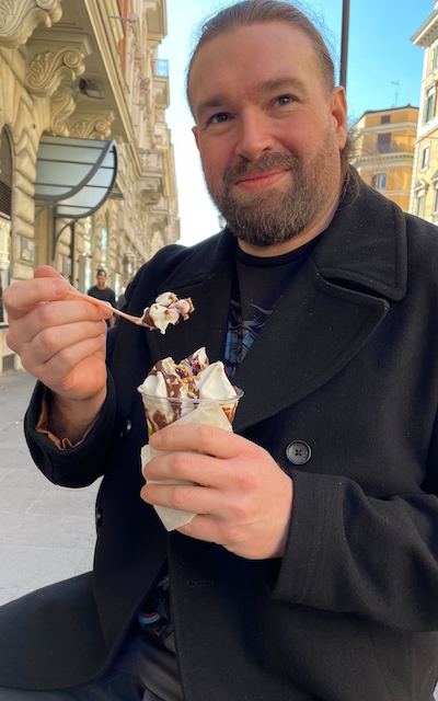 Ian eating a gelato in Rome, looking very content smiling at the camera
