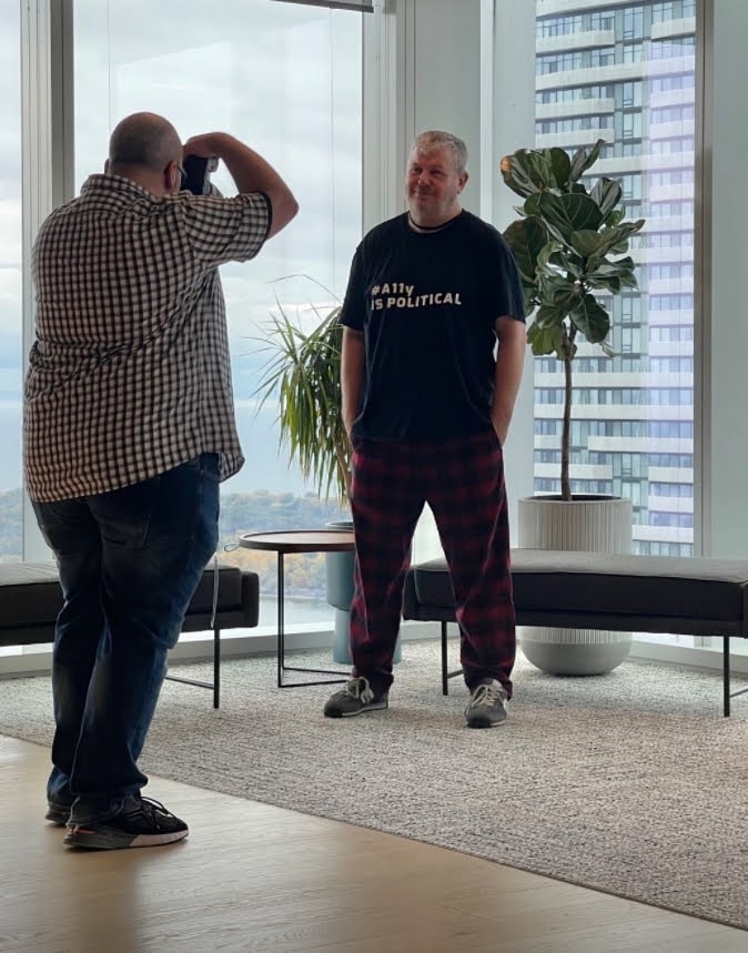 Steve in a full length photo wearing a t-shirt with "A11y is political" emblazoned on the front. Patrick is taking his picture with skyscrapers dominating the background