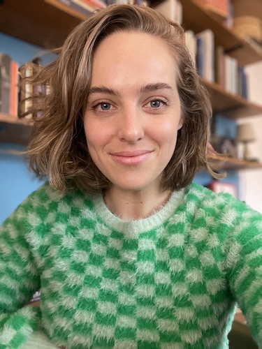 Demelza, smiling naturally at the camera, her brown hair flowing onto a jazzy, chequered, fluffy green jumper