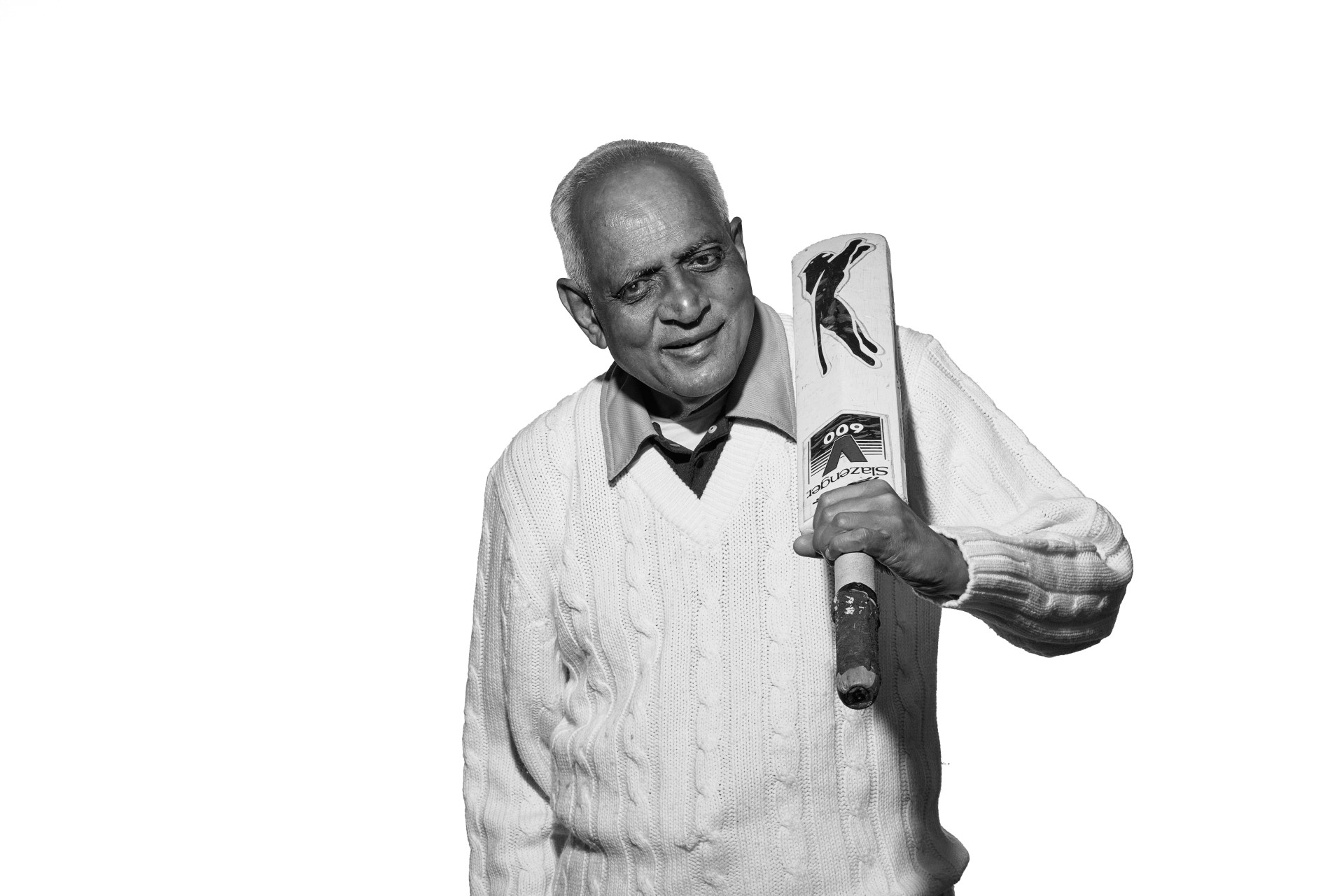 A black and white photo of Hasmukh, an older man of South Asian heritage. He is wearing his cricket whites smiling as he stands ready to bat