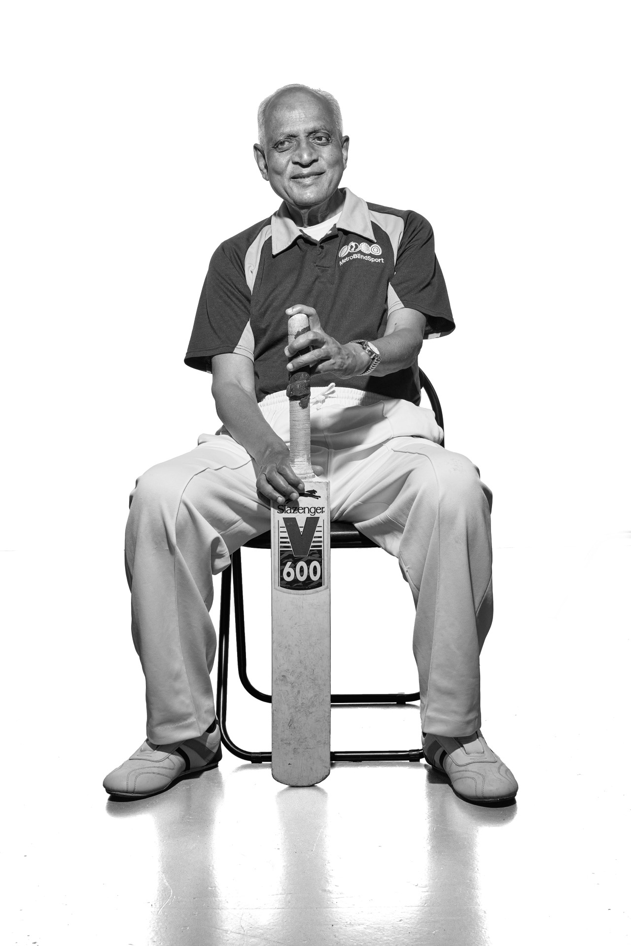 A black and white photo of Hasmukh, an older man of South Asian heritage. He is wearing his Metro Blind Sport cricket club shirt, sitting and proudly holding the top of his bat with both hands as the bottom rests on the floor as if he is waiting to go into bat