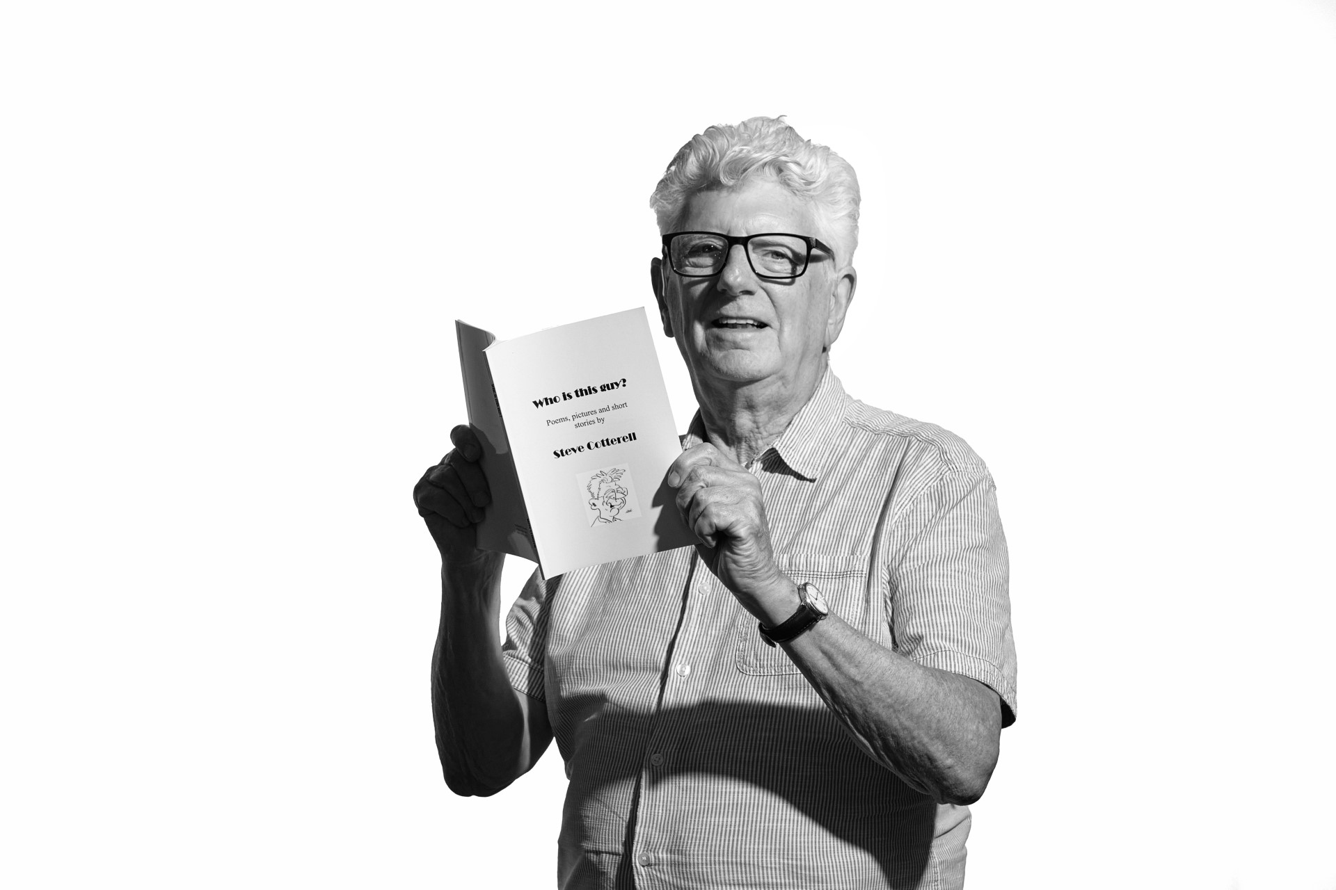 A black and white photo of Steve, an older white man with thick white hair. He is standing facing into the camera while he holds his book or poetry and photos open in front of him