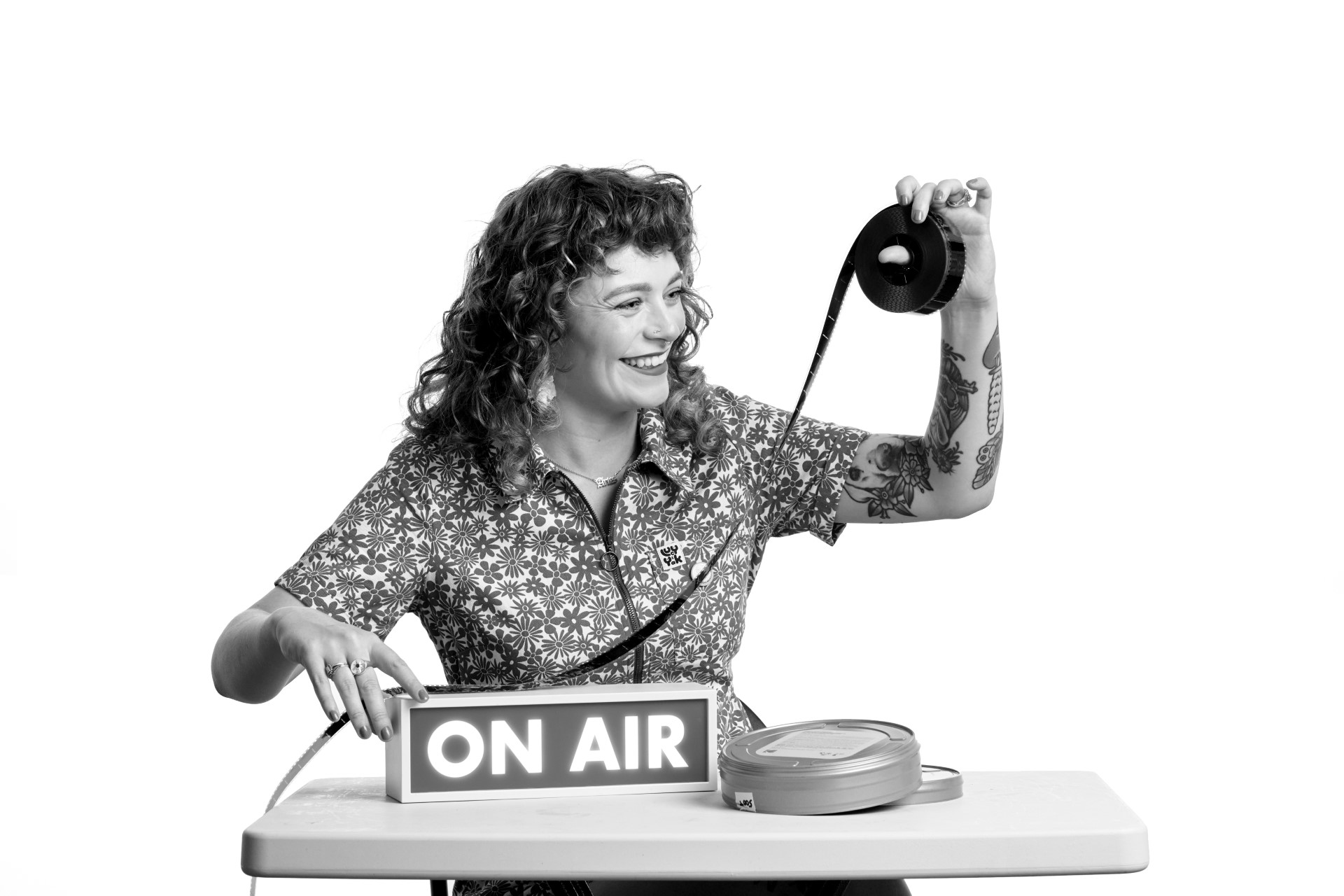 A black and white photo of Lauren, a younger white woman with long curly red hair. She is grinning as she sits at a desk with an On Air sign on it as she holds a strip of film up to the light with her left hand