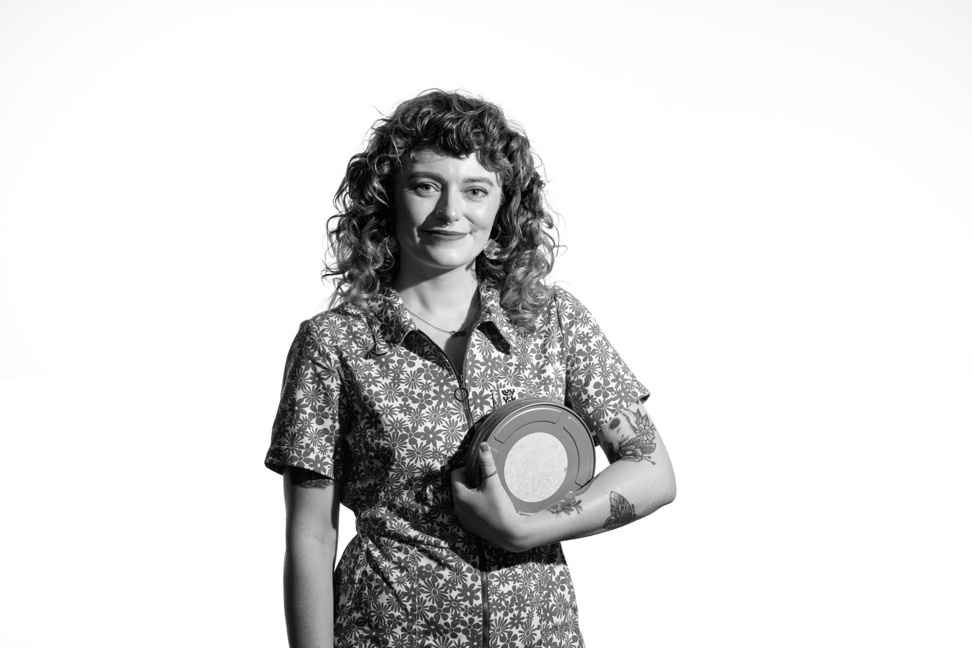 A black and white photo of Lauren, a younger white woman with long curly red hair. She is standing looking into the camera with a smile as she holds a flat round metal film canister in her left hand