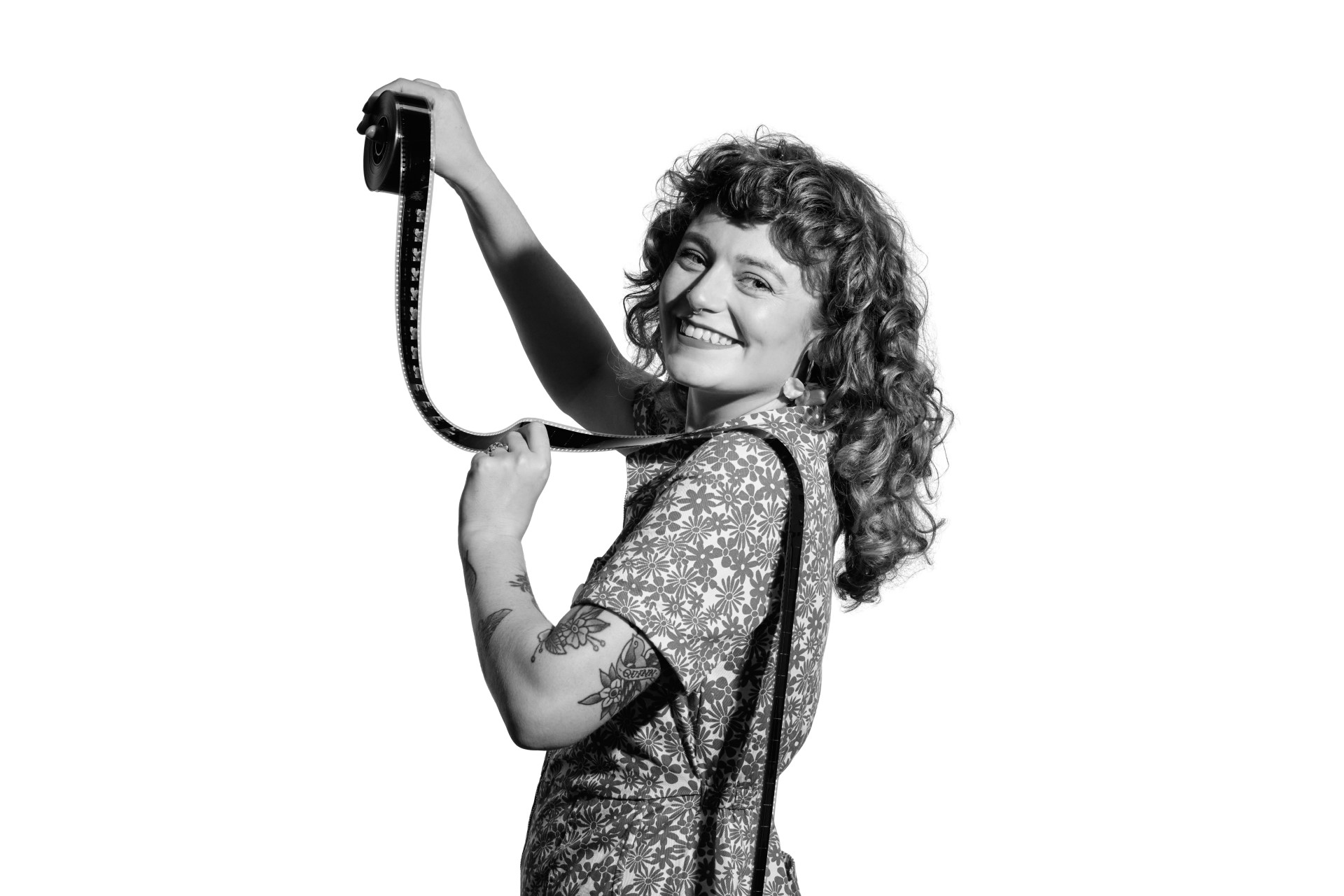 A black and white photo of Lauren, a younger white woman with long curly red hair. She is standing looking over her left shoulder into the camera with a smile as she holds a film reel up to the light with her right hand