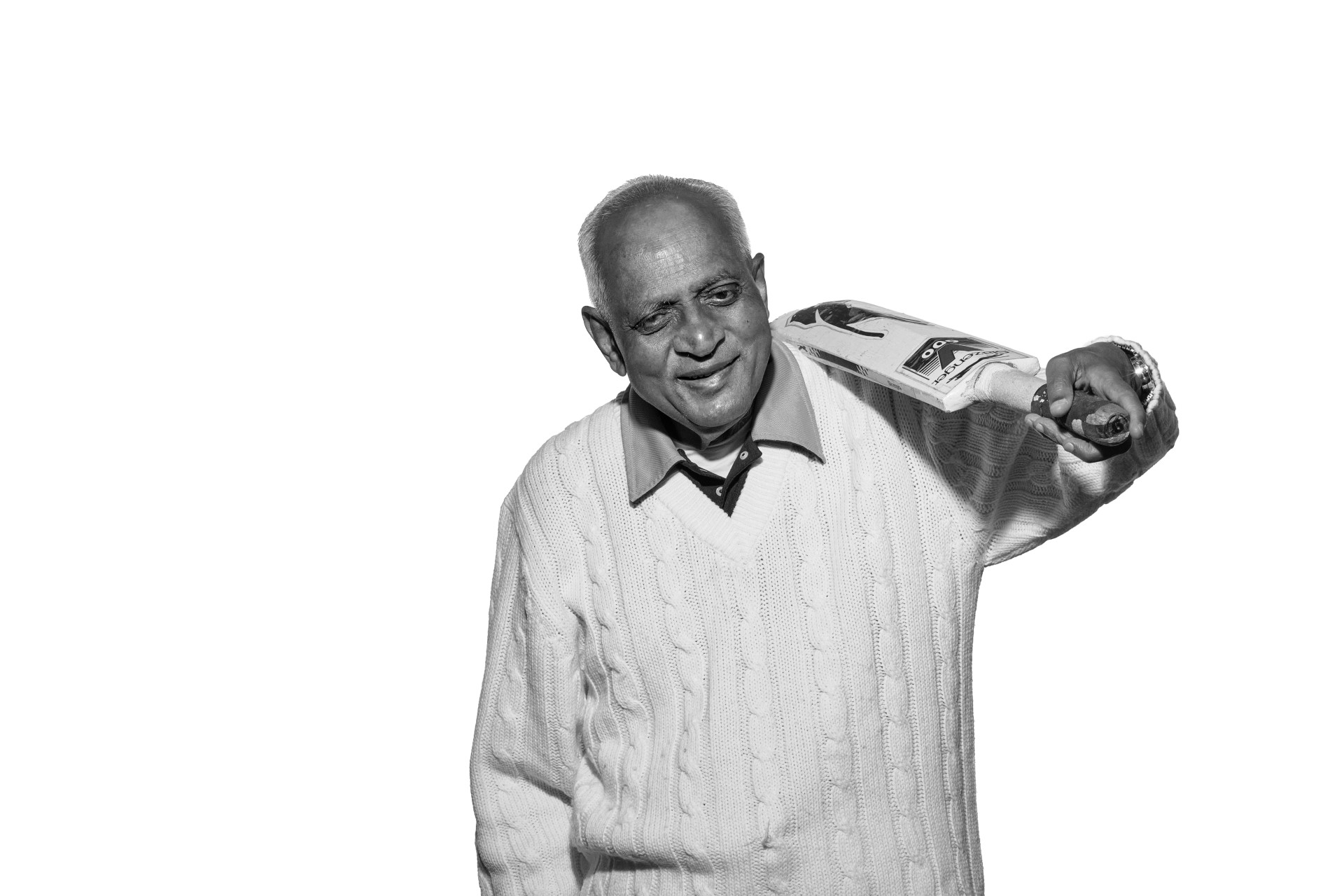 A black and white photo of Hasmukh, an older man of South Asian heritage. He is wearing his cricket whites standing and looking into the camera with a cheeky smile as he holds his bat which is resting on his left shoulder