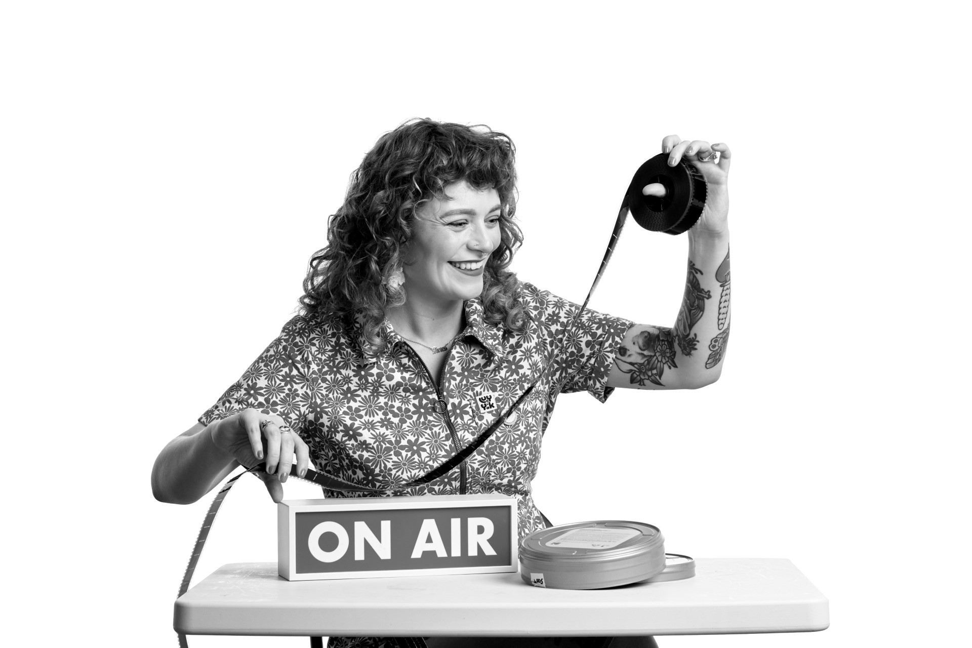 A black and white photo of Lauren, a younger white woman with long curly red hair. She is grinning as she sits at a desk with an On Air sign on it as she holds a strip of film up to the light with her left hand and inspects it closely