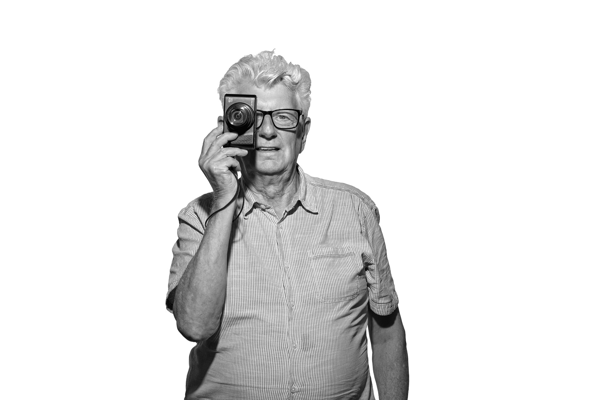 A black and white photo of Steve, an older white man with thick white hair. He is facing into the camera while he holds his camera over his right eye