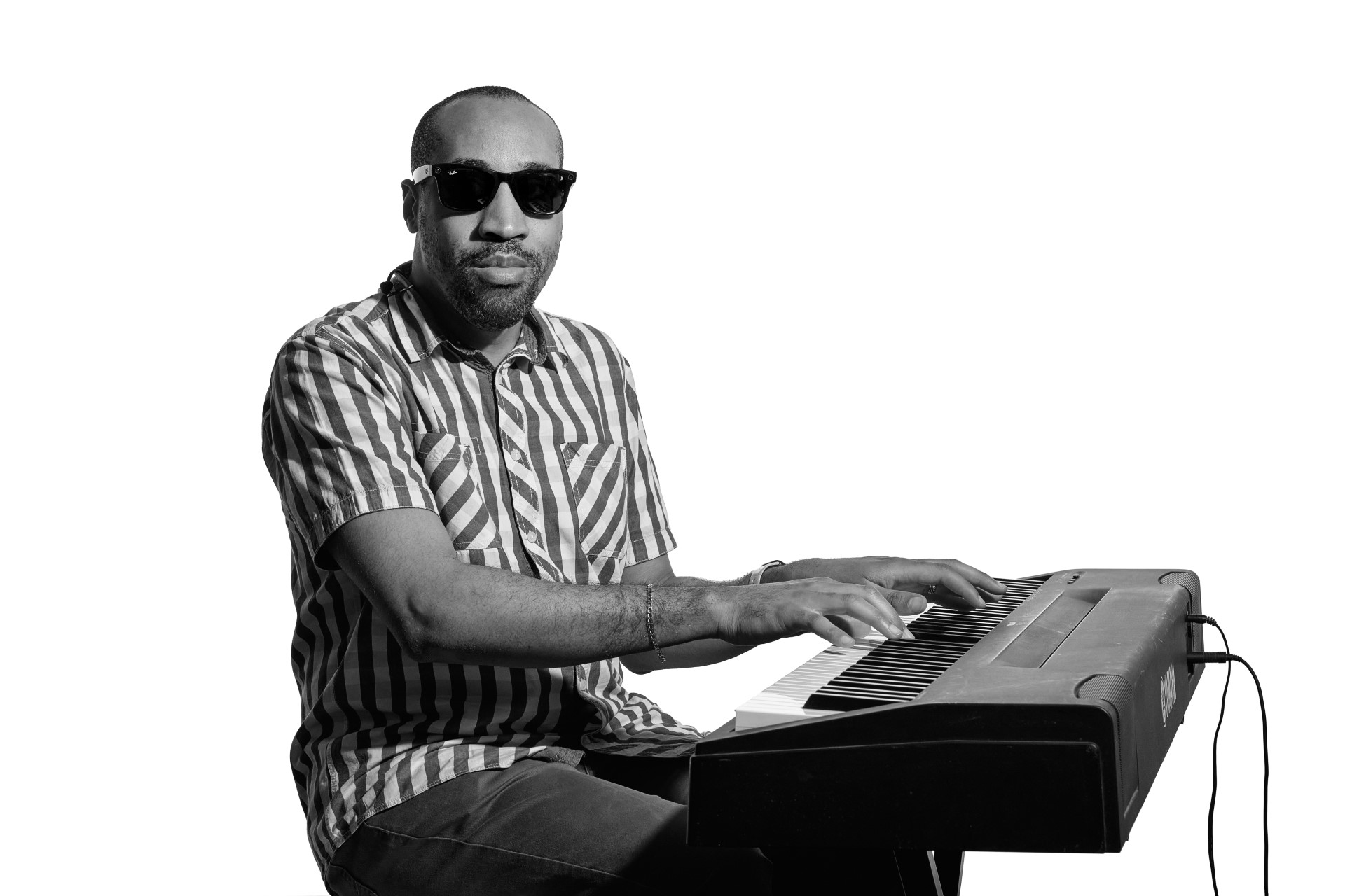 A black and white photo of Andre playing an electronic keyboard sitting side on facing the camera with a slight smile. Andre is a younger black man and wears a striped shirt and Ray-Ban Meta Smart Glasses