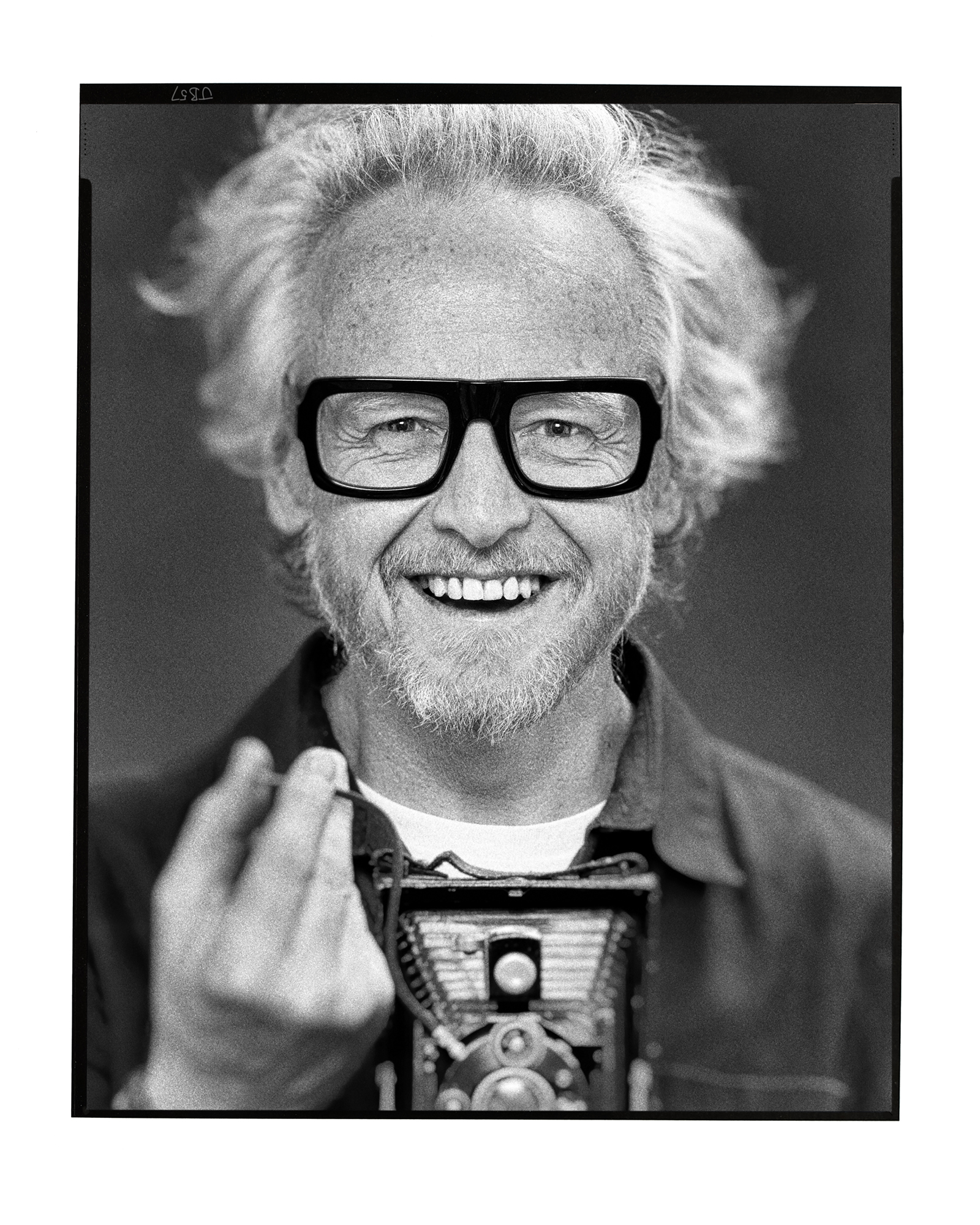 A black and white portrait of a man with wavy white hair, a short beard, and thick black glasses. He is smiling warmly and holding a vintage camera in front of him, with his right hand raised as he takes the picture using a button on the end of a cord. The background is blurred with a classic film border at the edges.