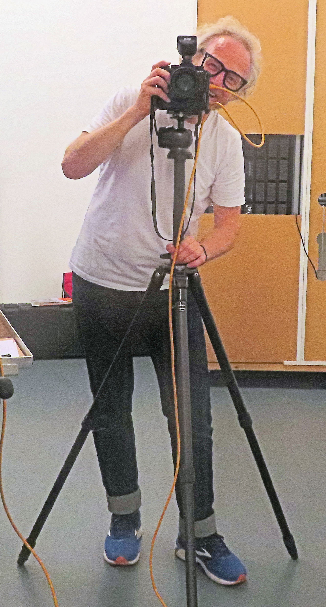 A photo of Jonathan, a man with white hair and thick black-framed glasses, standing in a studio behind a camera mounted on a tripod. He is wearing a white t-shirt, dark jeans with rolled-up cuffs, and blue sneakers with red soles. He is slightly leaning to one side, smiling, and looking through the camera’s viewfinder.