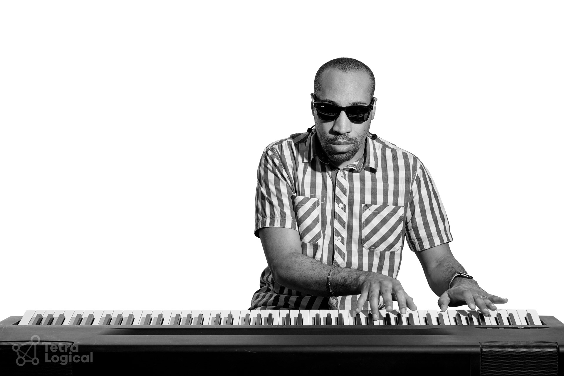 A black and white photo of Andre playing an electronic keyboard sitting face on looking down at his hands as he plays. Andre is a younger black man and wears a striped shirt and Ray-Ban Meta Smart Glasses