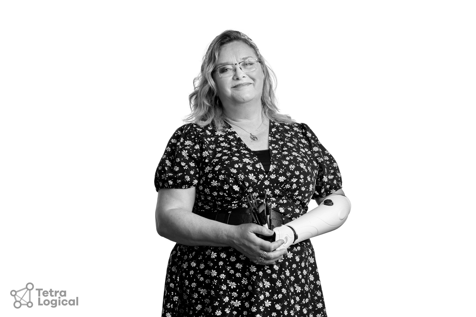 A black and white photo of Bev, a white woman with blond hair in her 50s scooped back in a hairband. She is standing looking into the camera, her head is cocked to the side with a smile while her hands are clasped together in front of her as she holds her brushes in her prosthetic hand and arm