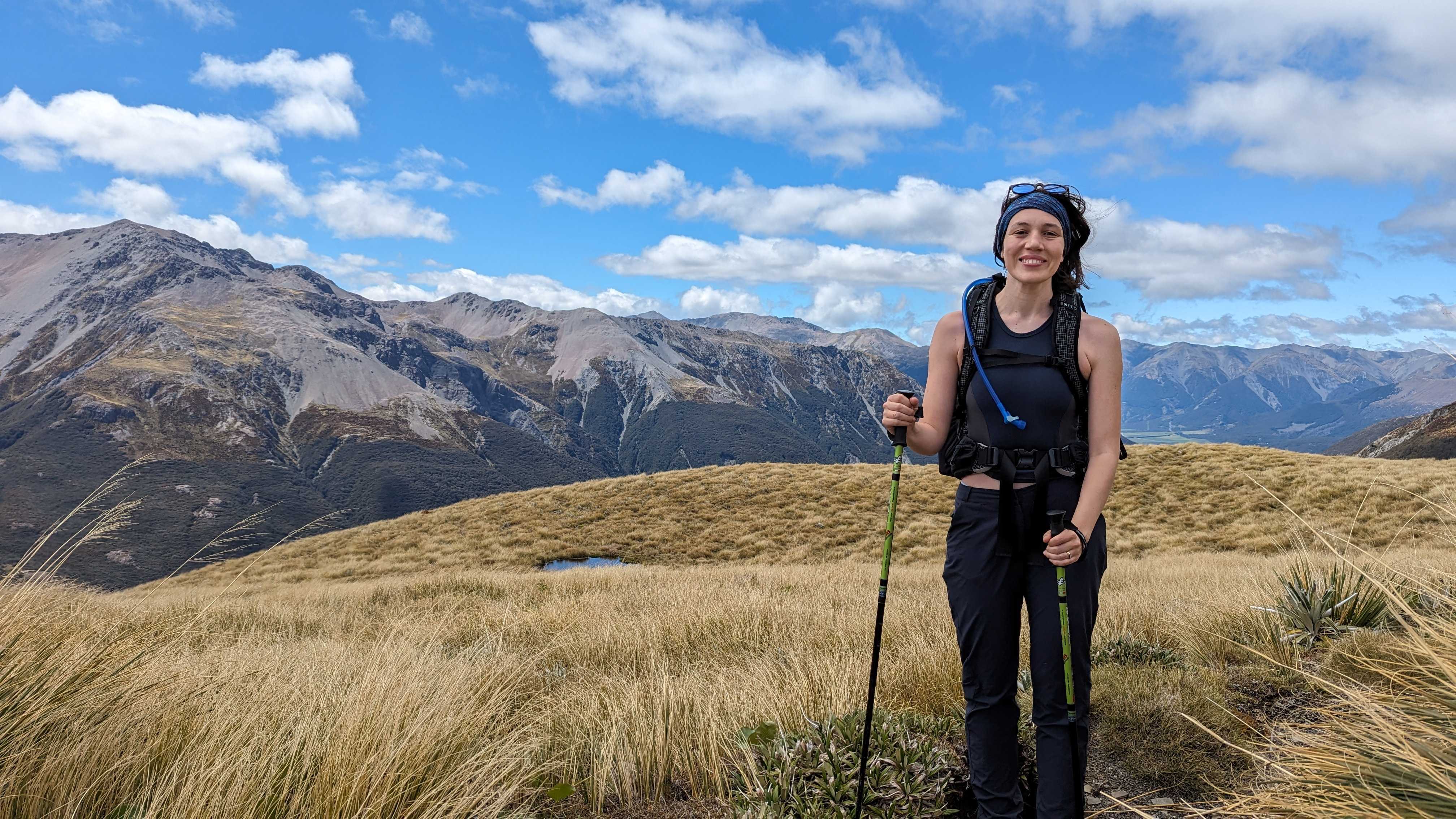 A vast landscape of rocky mountains sits underneath a blue, cloudy sky in New Zealand. Catriona stands in the foreground amid a sea of long grass, decked up in dark walking gear, holding a walking pole in either hand