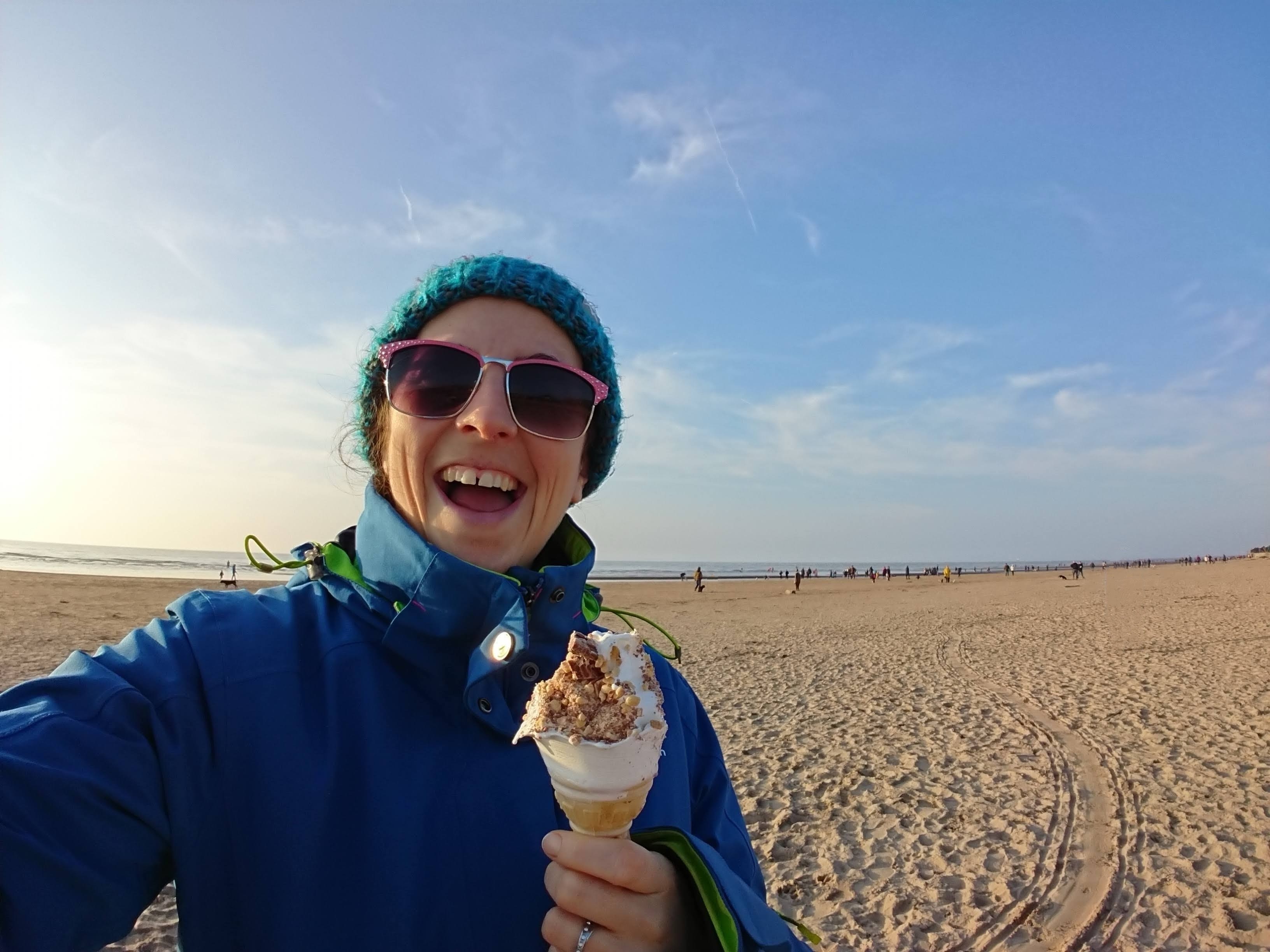 Grace stands on a sandy beach clad in a blue woolly hat and blue winter coat. She has pink sunglasses on, a huge ice cream in her hand and is beaming at the camera