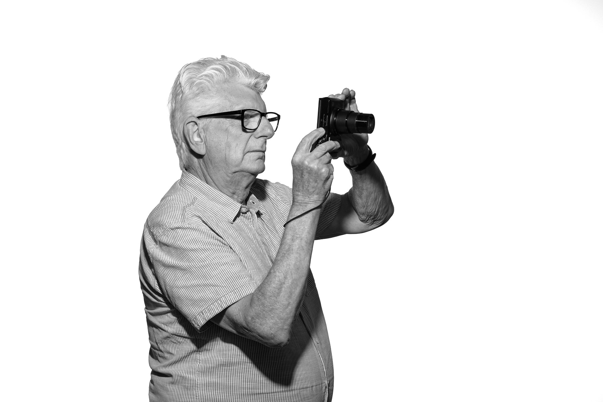 A black and white photo of Steve, an older white man with thick white hair. He is standing side on as he holds his camera up to his face looking focused
