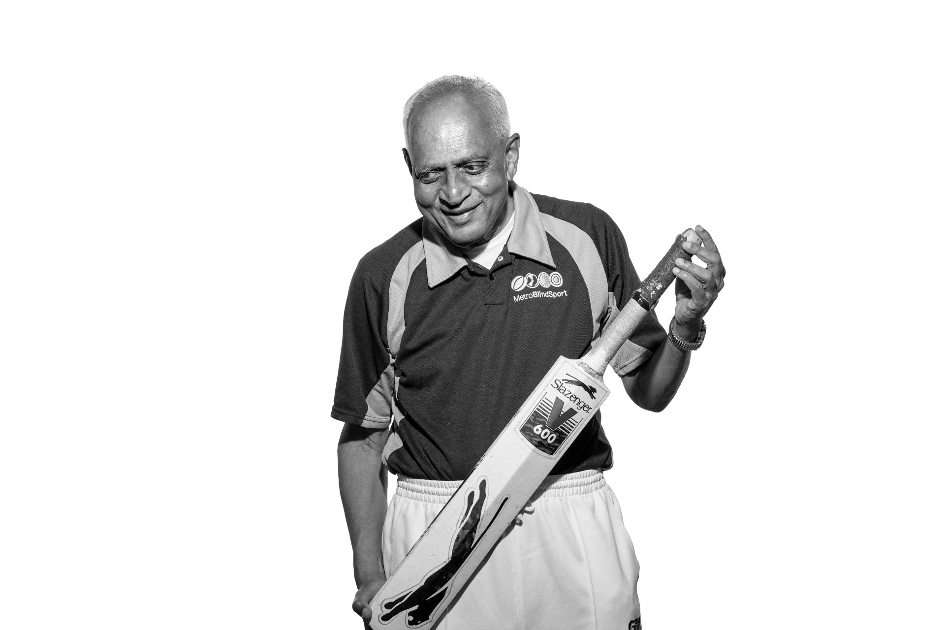 A black and white photo of Hasmukh an older man of South Asian heritage. He is wearing his Metro Blind Sport cricket club shirt, smiling as he holds his bat at the top with his left hand and bottom with his right across the front of his body